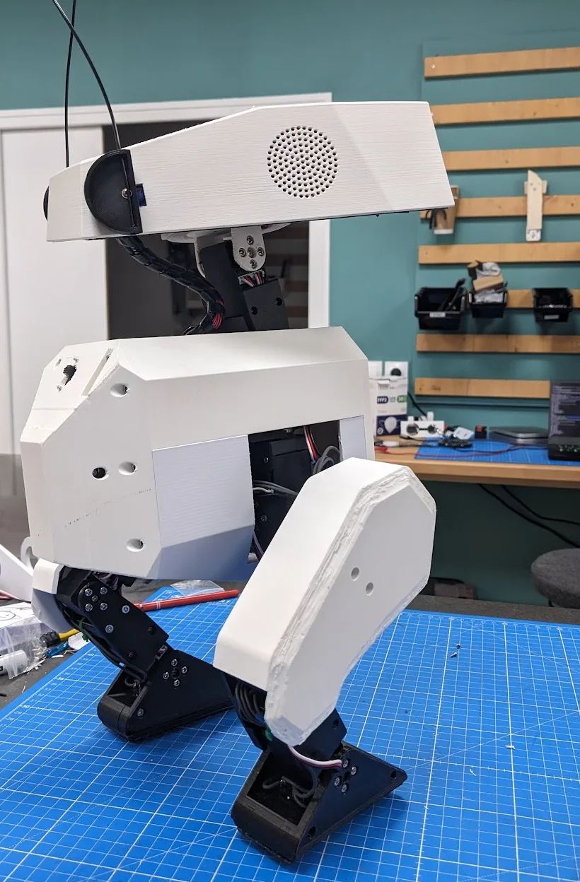 Open Duck Mini v2 on a workbench showing its printed body, exposed joints, wiring, and speaker opening
